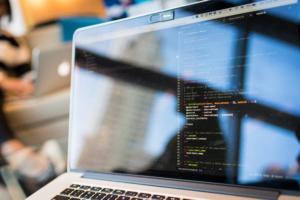 Eine Nahaufnahme eines Laptop-Bildschirms, der lebendigen Code in einer Programmierumgebung zeigt, mit Blick auf Ergonomie am Arbeitsplatz. Der unscharfe Hintergrund deutet auf eine weitere Person hin, die an einem Laptop arbeitet, und vermittelt so den Eindruck eines kollaborativen oder Coworking-Raums.