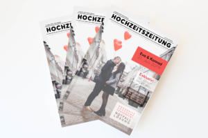 Drei Hochzeitsmagazine mit dem Titel „HOCHZEITSZEITUNG“ zeigen ein sich auf der Straße küssendes Paar mit roten herzförmigen Luftballons in den Händen. Das Cover mit Ideen zur Personalisierung Ihrer eigenen Hochzeitszeitung zeigt eine Hochzeit am „27. Juli 2019“, alles vor einem strahlend weißen Hintergrund.