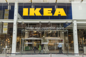 Das Bild zeigt die Fassade eines IKEA-Geschäfts, das mit einem großen gelben Logo sein Alleinstellungsmerkmal hervorhebt. Darunter stehen Glastüren mit der Aufschrift „Bestell- und Abholpunkt“. Drinnen sind verschiedene Einrichtungsgegenstände zu sehen, während sich ein paar Leute in der Nähe des geschäftigen Eingangs versammeln.