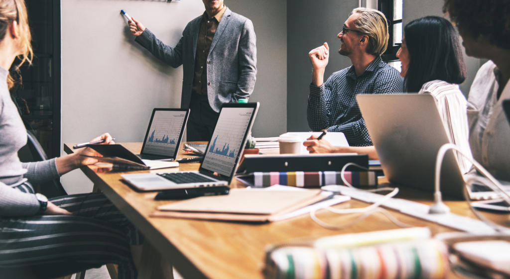 Bei einem Meeting mit fünf Personen steht eine Person und zeigt auf ein Whiteboard, um das Alleinstellungsmerkmal hervorzuheben, während die anderen mit Laptops und Dokumenten um einen Tisch sitzen. Sie wirken konzentriert und in die Diskussion vertieft.