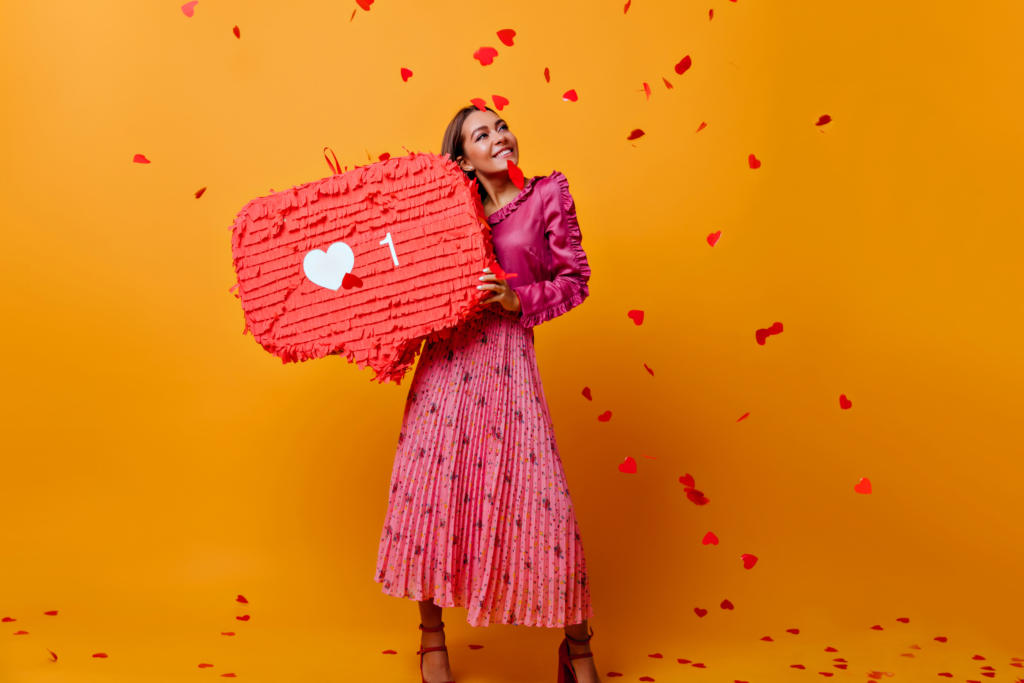 Eine Frau in rosa Bluse und Faltenrock lächelt und hält eine große rote herzförmige Piñata mit einem weißen Herzsymbol und der Zahl 1 in der Hand, die subtil ihr Alleinstellungsmerkmal zur Schau stellt. Kleines rotes Herzkonfetti fällt vor einem orangefarbenen Hintergrund um sie herum herab.