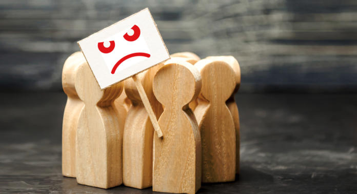 Holzfiguren, die Menschen darstellen, die zusammenkommen, symbolisieren den Gemeinschaftsgeist. Eine Figur hält ein Schild mit einem roten, wütenden Gesicht und weist auf die Herausforderungen hin, die die Aufrechterhaltung der Kundenbindung vor einem dunklen, strukturierten Hintergrund mit sich bringt.