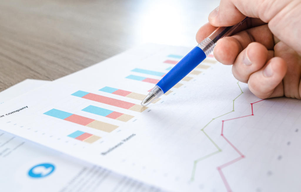 Nahaufnahme einer Hand, die einen blauen Stift hält und auf gedruckte Diagramme und Grafiken zeigt. Die für ein effektives Beschwerdemanagement wichtigen Dokumente zeigen farbenfrohe Balken- und Liniendiagramme mit Finanz- oder Geschäftsdaten auf einem Holztisch.