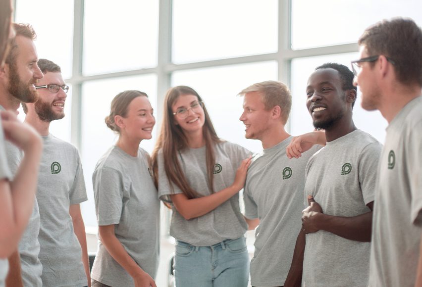 Einige Teamkolleg*innen stehen lächelnd beisammen und tragen alle das gleiche graue Shirt mit Firmenlogo Einige Teamkolleg*innen stehen lächelnd beisammen und tragen alle das gleiche graue Shirt mit Firmenlogo