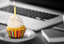 Ein Cupcake mit weißem Zuckerguss und einer brennenden gelben Kerze steht auf einem weißen Teller. Der Cupcake hat eine bunt gestreifte Verpackung. Im Hintergrund liegen ein Laptop, ein Notizbuch und ein Stift auf einem grauen Schreibtisch.