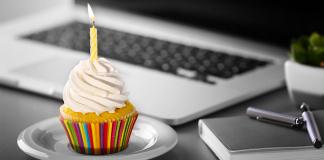 Ein Cupcake mit weißem Zuckerguss und einer brennenden gelben Kerze steht auf einem weißen Teller. Der Cupcake hat eine bunt gestreifte Verpackung. Im Hintergrund liegen ein Laptop, ein Notizbuch und ein Stift auf einem grauen Schreibtisch.