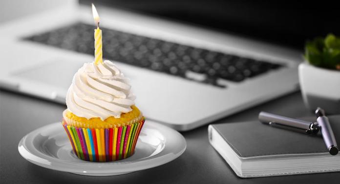 Ein Cupcake mit weißem Zuckerguss und einer brennenden gelben Kerze steht auf einem weißen Teller. Der Cupcake hat eine bunt gestreifte Verpackung. Im Hintergrund liegen ein Laptop, ein Notizbuch und ein Stift auf einem grauen Schreibtisch.