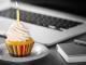 Ein Cupcake mit weißem Zuckerguss und einer brennenden gelben Kerze steht auf einem weißen Teller. Der Cupcake hat eine bunt gestreifte Verpackung. Im Hintergrund liegen ein Laptop, ein Notizbuch und ein Stift auf einem grauen Schreibtisch.